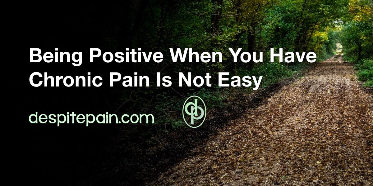 Being positive when you have chronic pain is not easy. A dart picture of a path leading through trees in a forest. Light is coming through the trees at the end of the path.